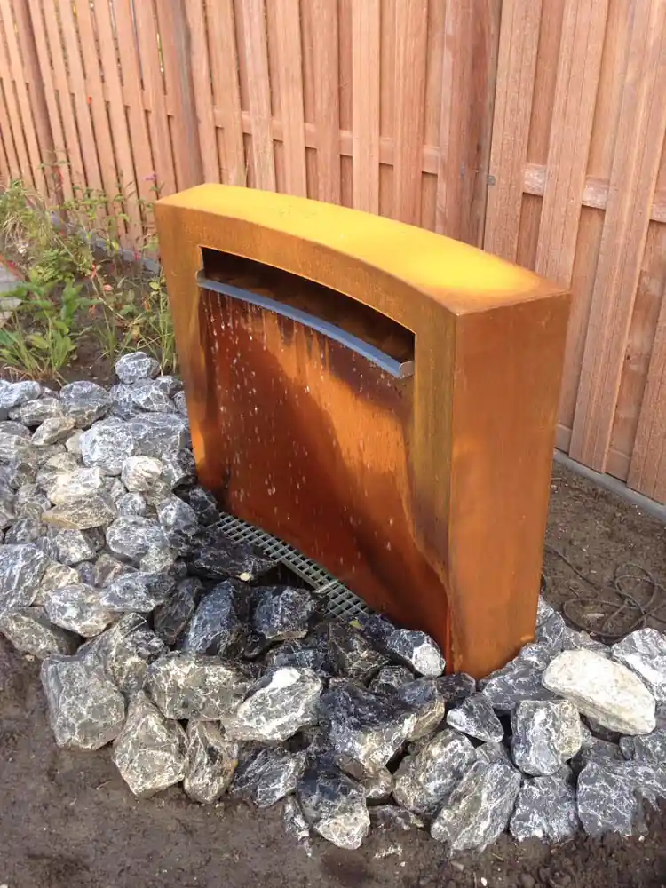 Cortenstalen waterwand met een brede waterval die uitstroomt over een bed van grijs gevlamde zwerfkeien, tegen een hardhouten schutting