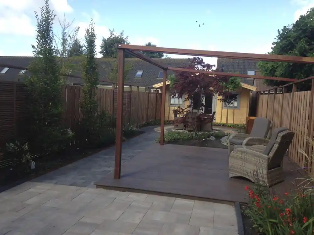 Overzicht van de achtertuin met een hardhouten pergola boven het vlonderterras, zuilvormige bomen langs de schutting en het tuinhuis met rond terras op de achtergrond