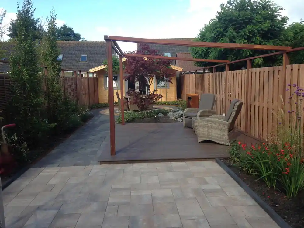 Hardhouten pergola boven een vlonder met rieten loungestoelen, met een klinkerpad langs de borders naar het ronde terras bij het tuinhuis