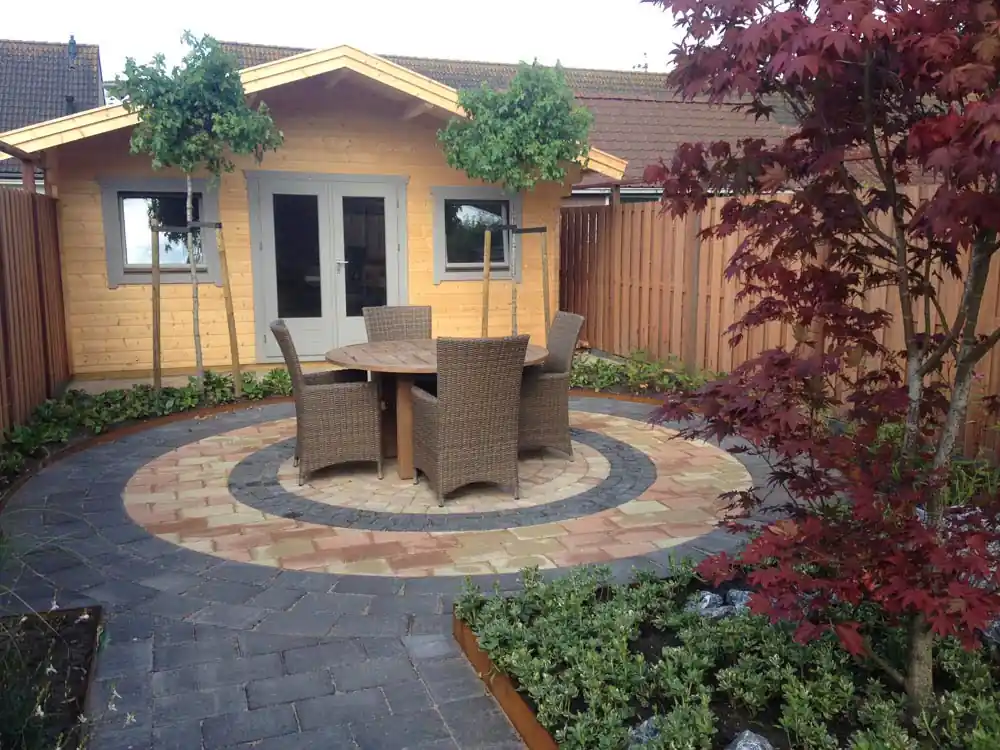 Rond terras van siermetselwerk in banen zandkleurige en antracieten klinkers met een ronde tuintafel en rieten stoelen, voor een houten tuinhuis met leibomen