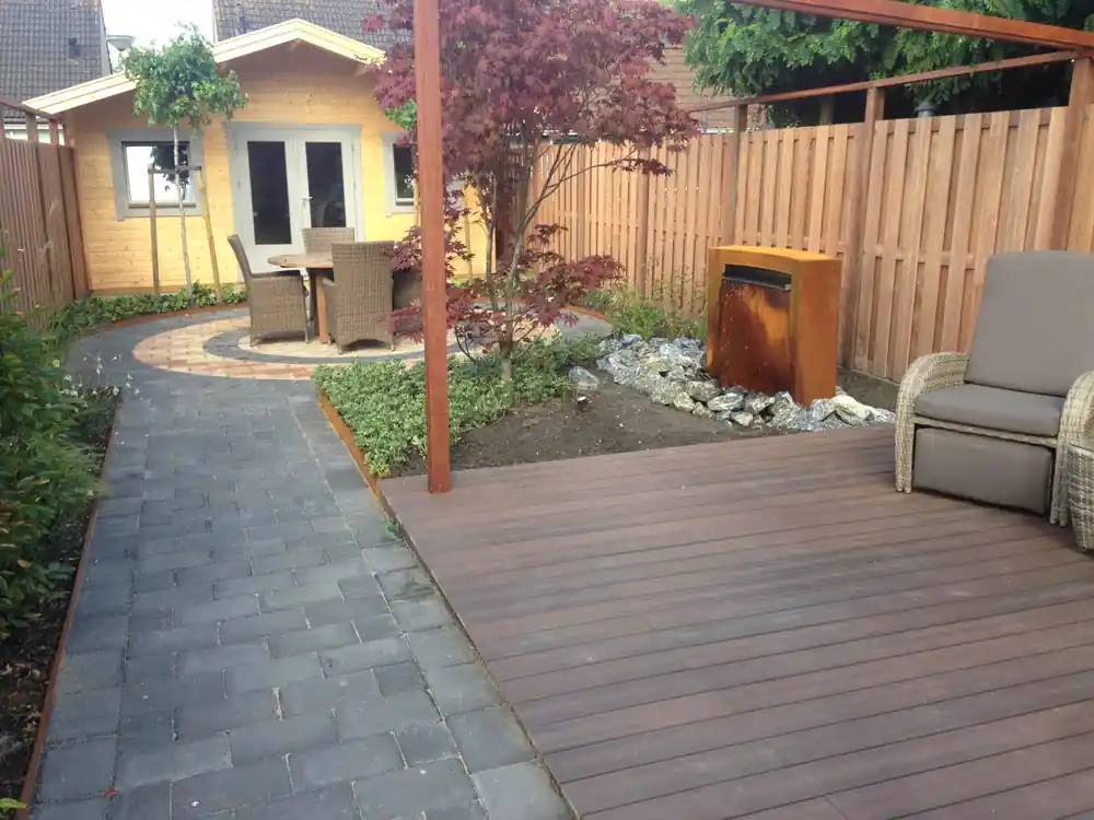 Hardhouten vlonder met loungestoelen onder een pergola, met een Japanse esdoorn, de cortenstalen waterwand en het ronde siermetselwerk terras erachter