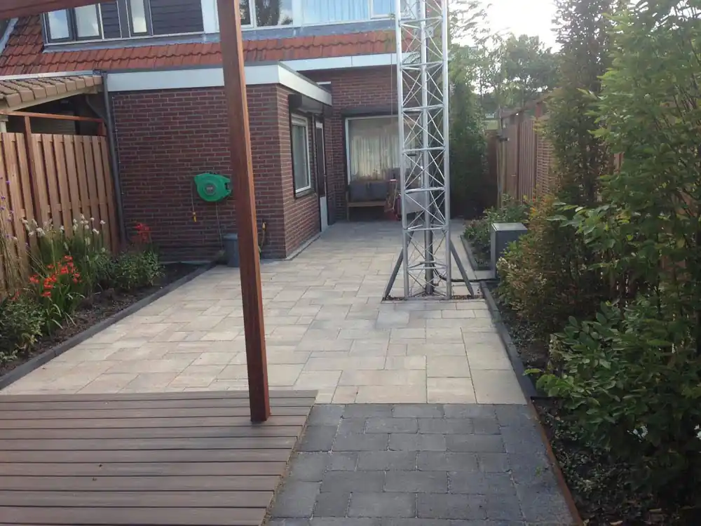 Bestraat zijpad langs de bakstenen woning met borders met montbretia en siergrassen, tijdens de aanleg met een steiger op het terras