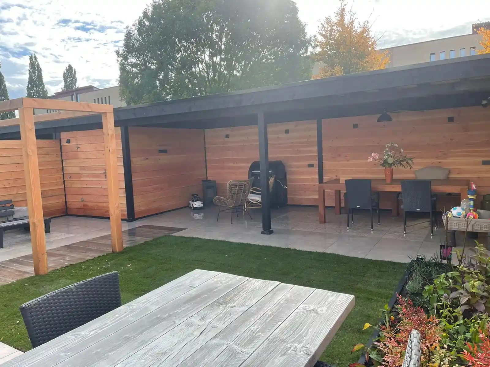 Ruime overkapping met Douglas wanden, een eettafel met zwarte stoelen en een aangebouwde berging, met een gazon en een houten picknicktafel op de voorgrond