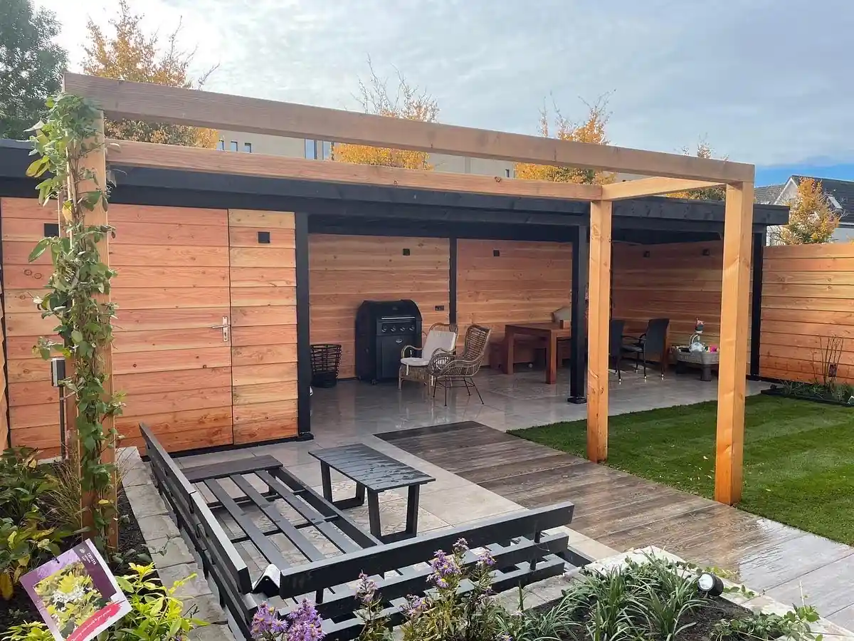Overkapping met Douglas wanden en bergingsdeur naast een pergola boven een vlonder met antracieten loungebank, met gazon, borders en een klimplant langs de staander