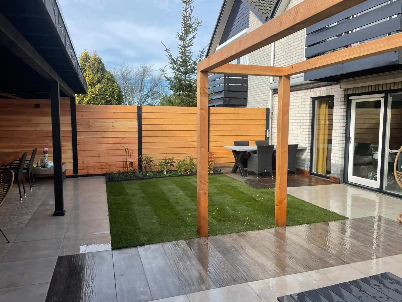 Douglas pergola boven een terras van lichte tegels met een strak gazon ernaast, met een eethoek bij de openslaande deuren en een border langs de Douglas schutting
