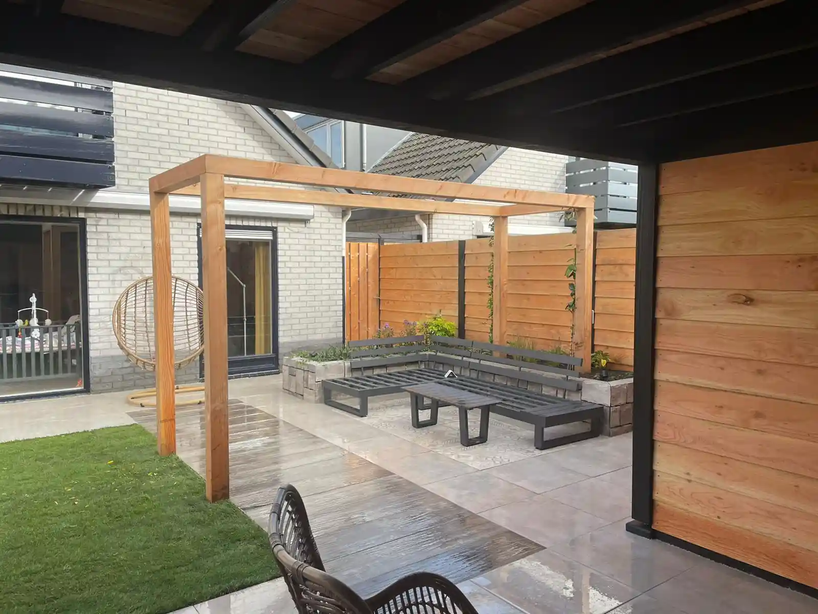 Douglas pergola boven een antracieten hoekbank tegen een verhoogde border van stapelblokken, met een vlak patroontegels, een hangstoel en een lichte gevel