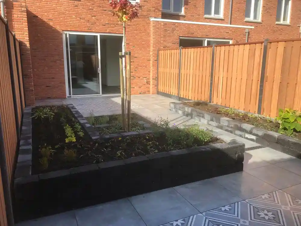 Ruime verhoogde border van zwarte stapelblokken met verse beplanting rond een jonge sierboom, met een tegelterras en de schuifpui op de achtergrond