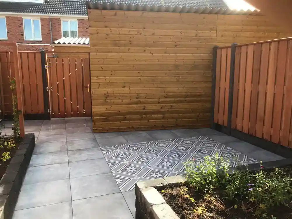 Houten bergingswand met daarnaast een poort in de schutting, met een terras van grijze tegels en een vlak witgrijze patroontegels ervoor