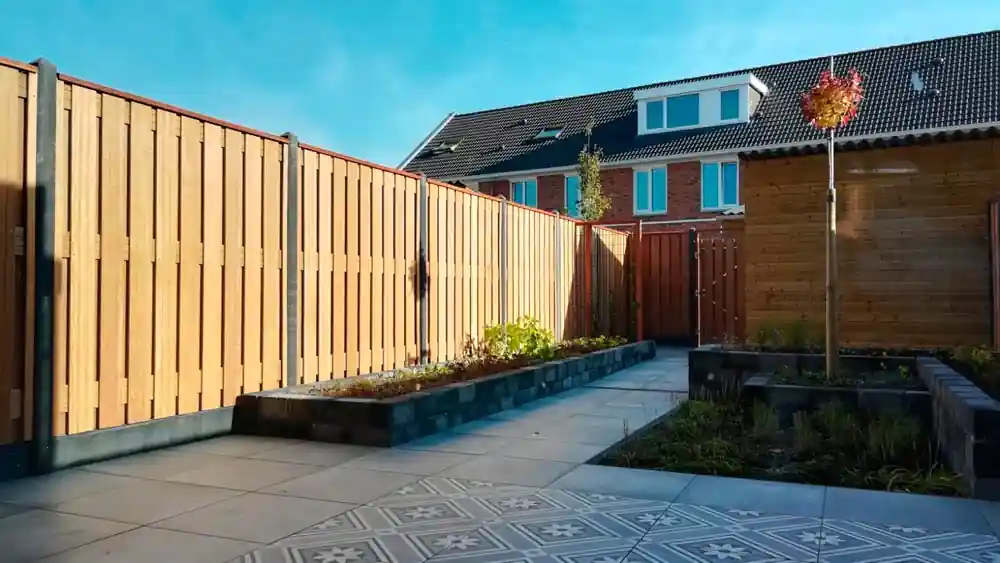 Smalle verhoogde border met jonge beplanting langs een hardhouten schutting, met een tegelpad en een vlak patroontegels op de voorgrond