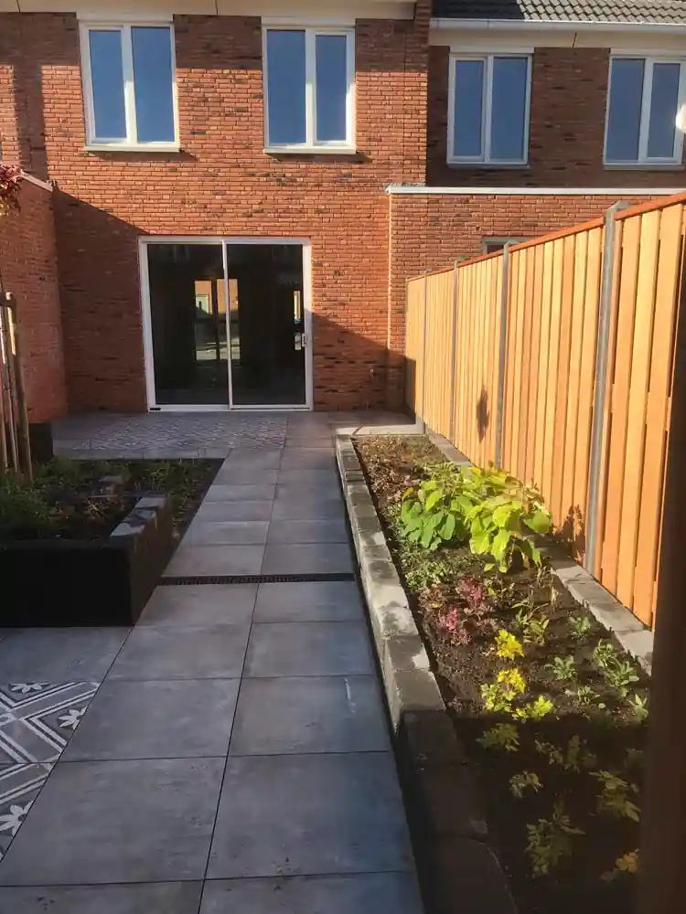 Tegelpad met lijngoot langs een border met verse beplanting, met zicht op de schuifpui van de bakstenen woning