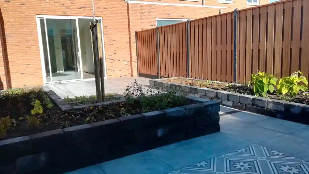 Verhoogde border van zwarte stapelblokken met verse beplanting, grenzend aan een terras met grijze tegels en een vlak met witgrijze patroontegels