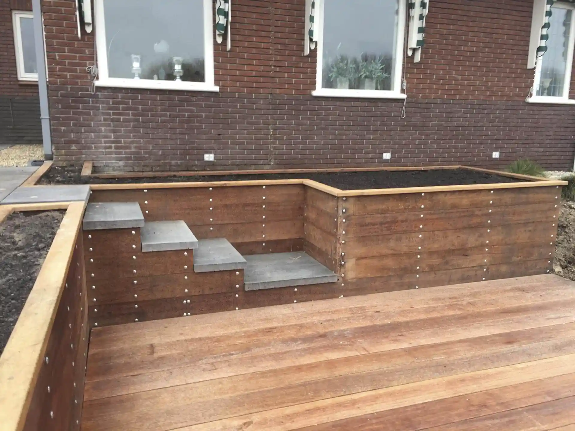 Verhoogde border met hardhouten keerwand en zichtbare bouten langs de gevel, met een trap van betonnen traptreden die afdaalt naar een hardhouten vlonder