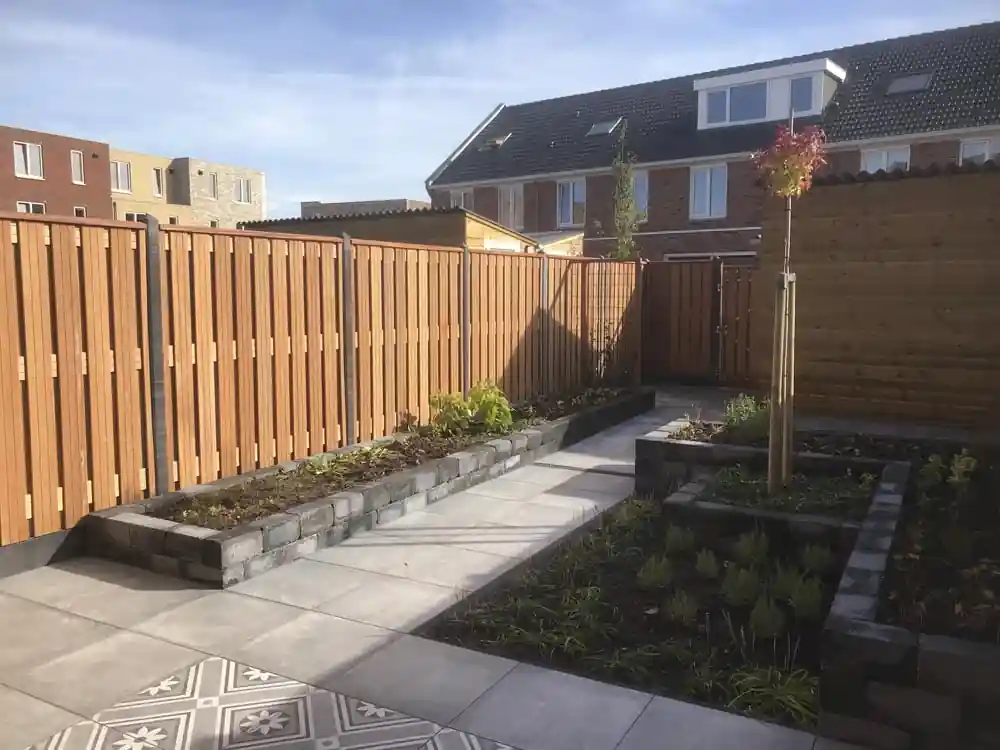 Langgerekte verhoogde border met jonge beplanting langs een hardhouten schutting, met een tegelpad dat naar een houten poort leidt