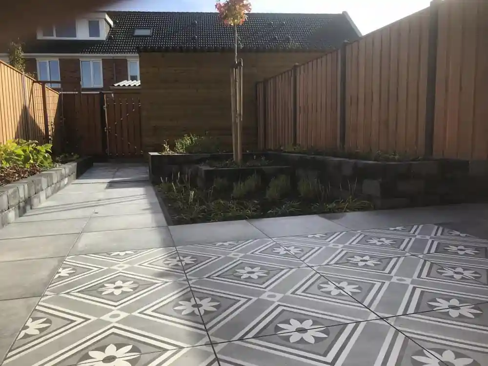 Verhoogde border van zwarte stapelblokken met lavendel en siergrassen voor een houten bergingswand, met een terras van patroontegels op de voorgrond