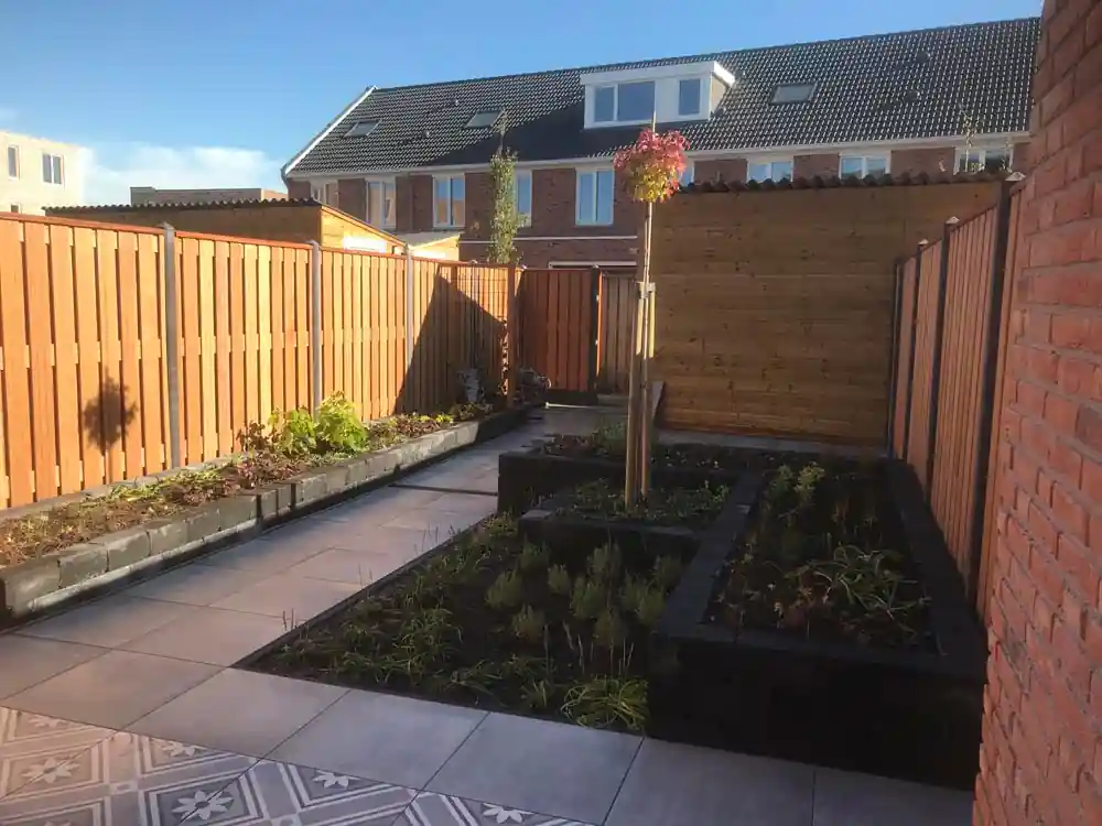 Achtertuin met een tegelpad tussen verhoogde borders met verse beplanting, met een houten poort, bergingswand en hardhouten schutting
