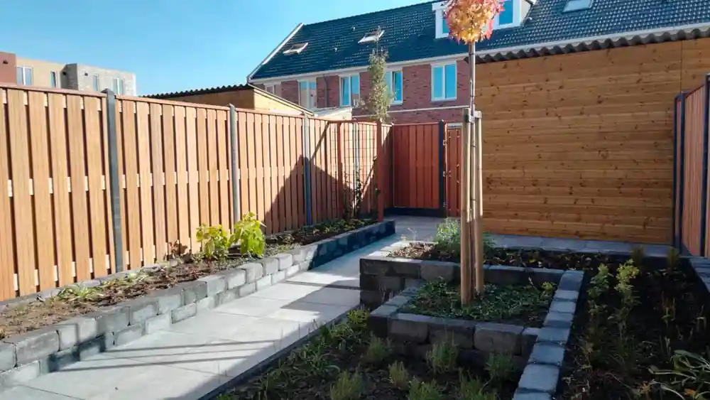 Tegelpad tussen twee verhoogde borders van grijze en zwarte stapelblokken, met een houten schutting, een poort en een jonge boom met boompaal