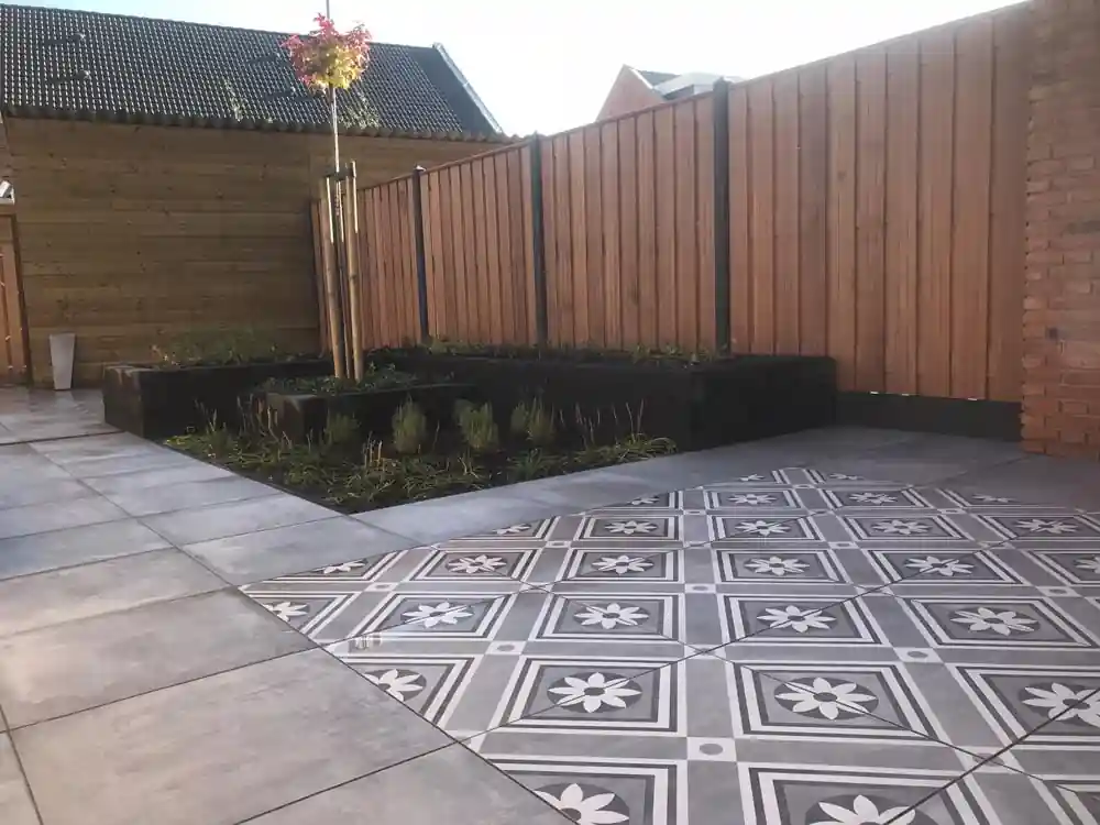 Vlak witgrijze patroontegels met bloemmotief naast grote grijze terrastegels, met een verhoogde border met lavendel langs de schutting