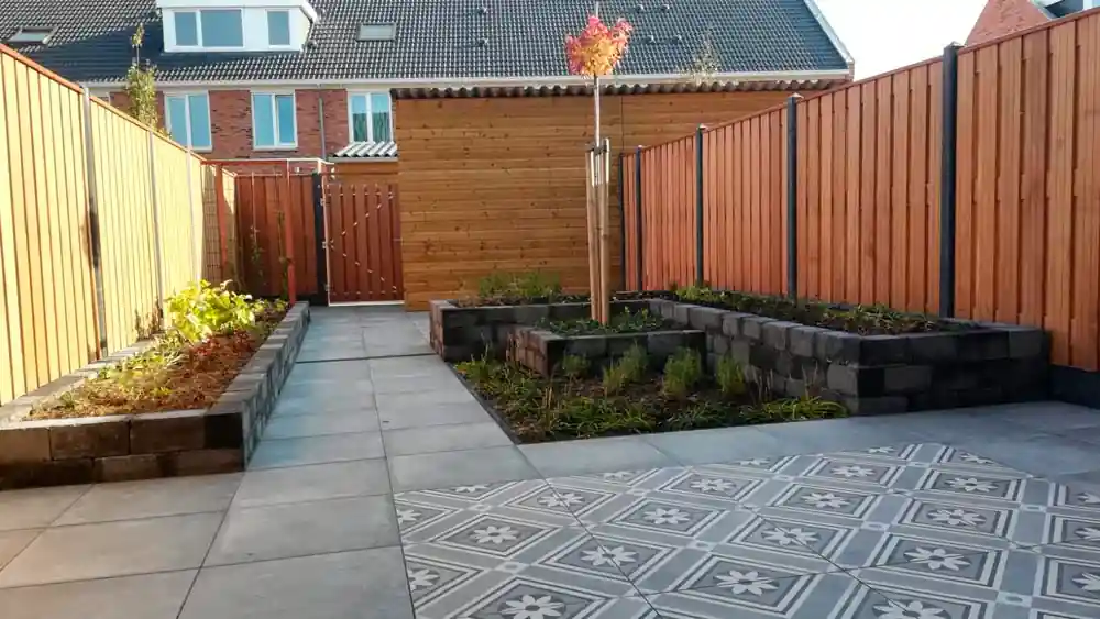 Terras van grijze tegels met een vlak sierlijke patroontegels, omringd door verhoogde borders van stapelblokken en een hardhouten schutting
