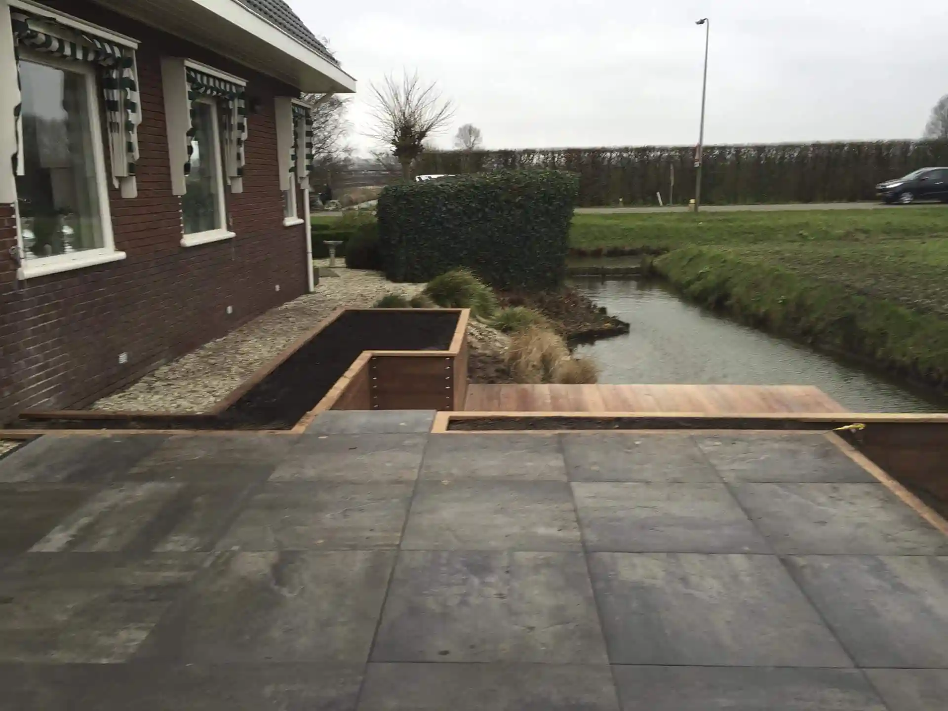 Hardhouten plantvakken met verse tuinaarde langs de bakstenen gevel en het tegelterras, met een beukenhaag, siergrassen en een vlonder aan het water
