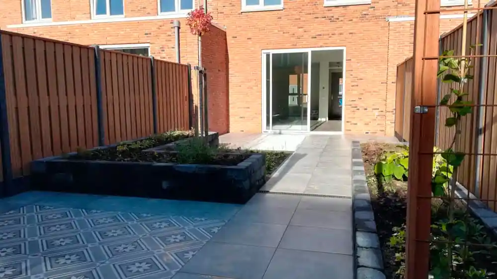Pad van grote grijze tegels naar de schuifpui van een bakstenen woning, met een verhoogde border links en een klimrek met jonge klimplant rechts