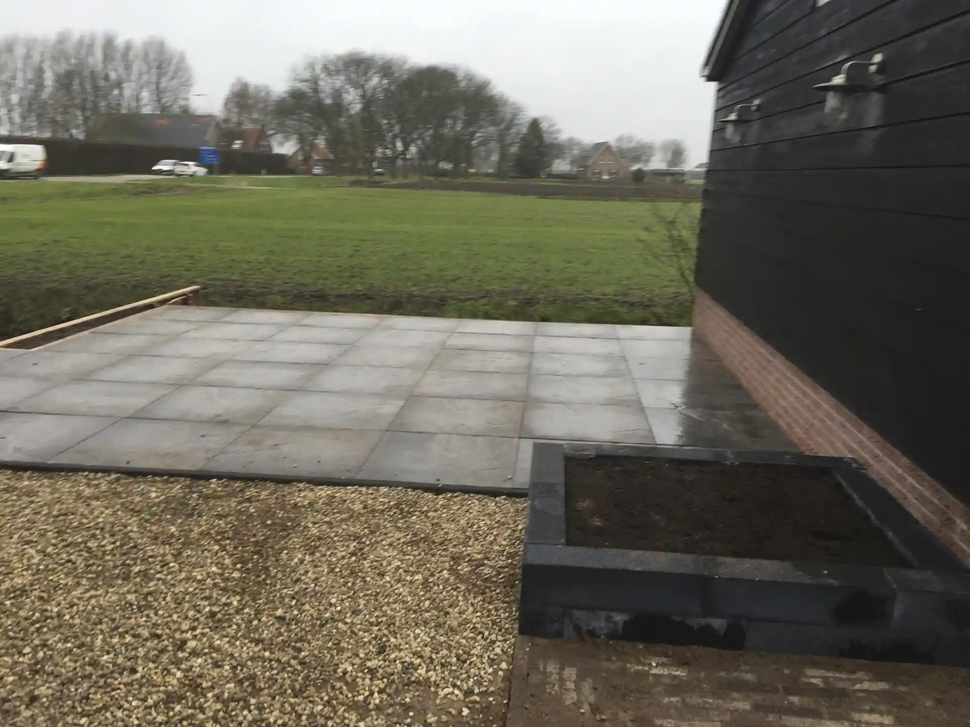 Nieuw aangelegd terras van grote grijze tegels tot aan de slootkant, met een strook split en een verhoogd plantvak van hardsteen langs de bakstenen plint