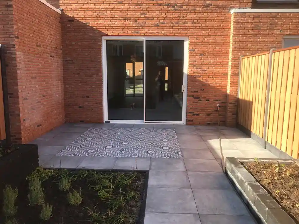 Terras van grijze tegels met een vierkant vlak witgrijze patroontegels voor een schuifpui, tussen een bakstenen muur en een lichte houten schutting