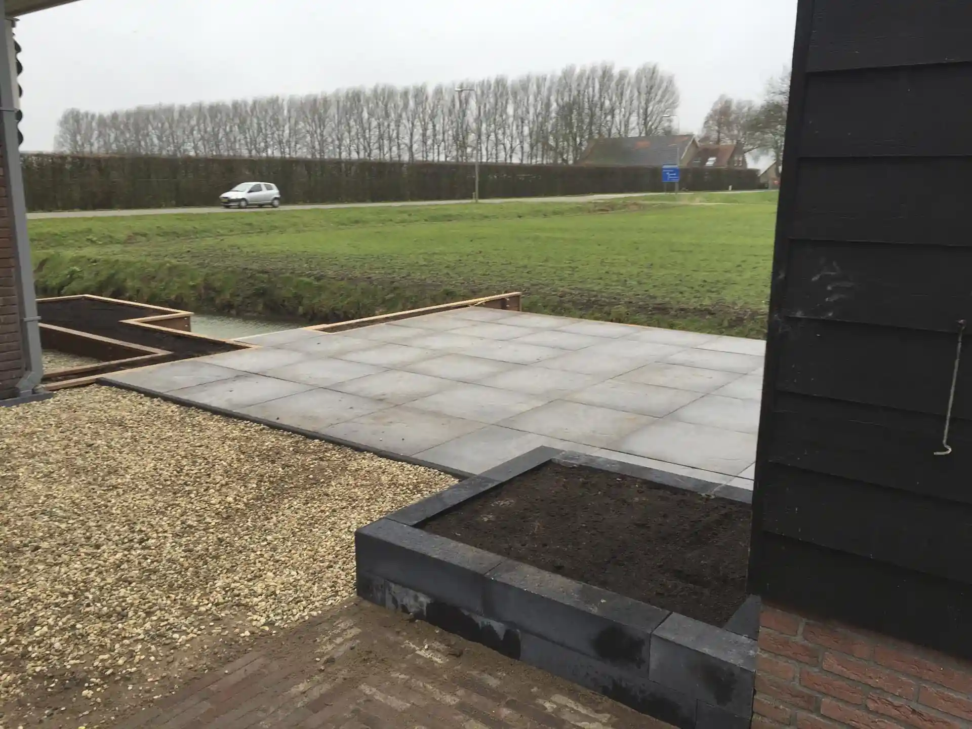 Terras van grote grijze tegels naast een vlak split, met een verhoogd plantvak van hardsteen langs de zwarte gevel en uitzicht over de sloot en het weiland