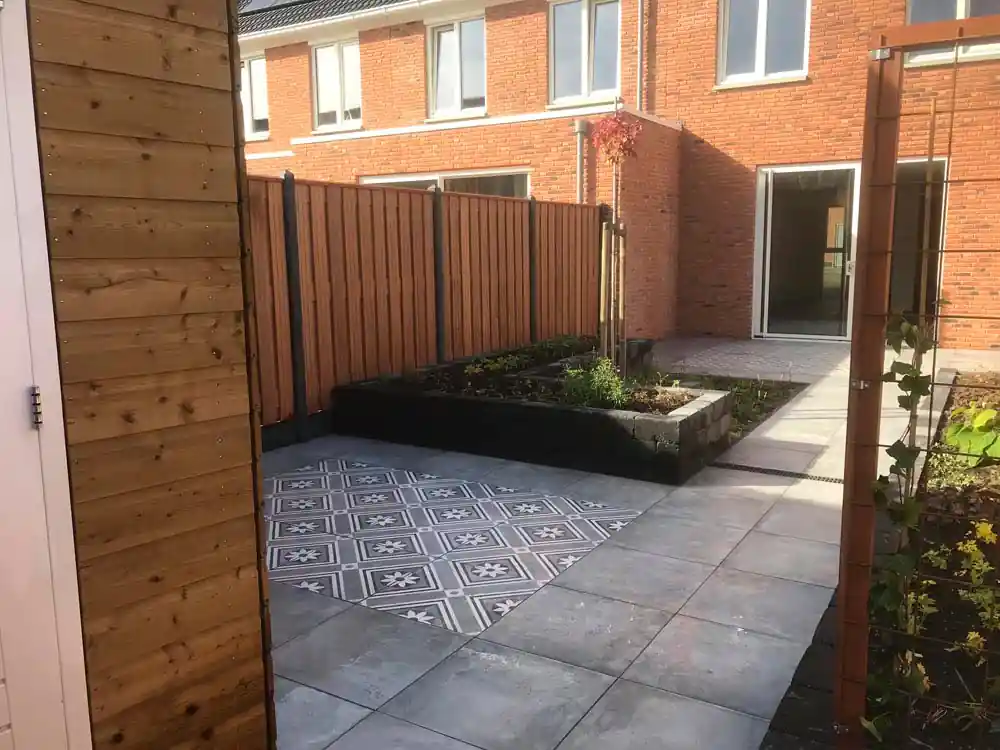 Terras van grijze tegels met een vlak patroontegels tussen een verhoogde border en een houten klimrek met jonge klimplant