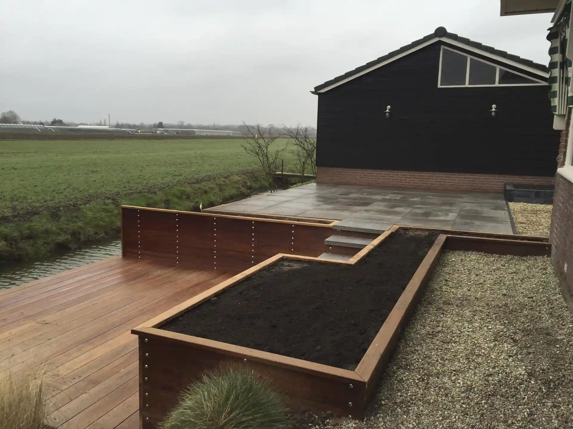 Hardhouten vlonder langs de slootkant met een keerwand en verhoogde plantvakken, met een tegelterras bij de zwarte schuurgevel en uitzicht over het weiland