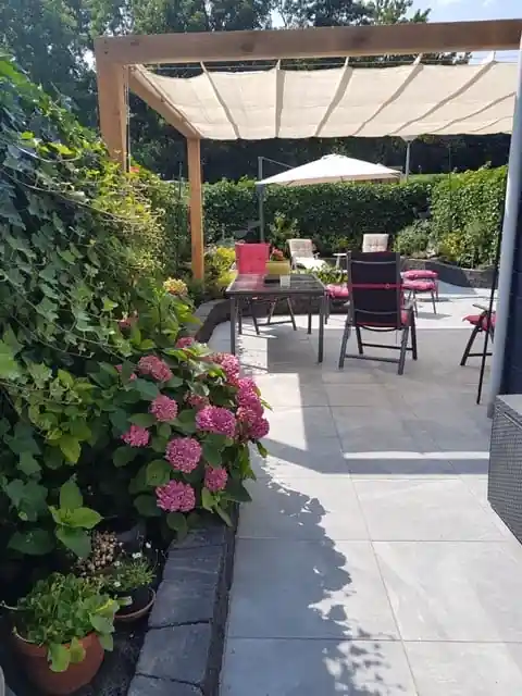 Houten pergola met cr?mekleurig schaduwdoek boven een eethoek, met op de voorgrond een border met roze hortensia's langs een terras van lichte tegels