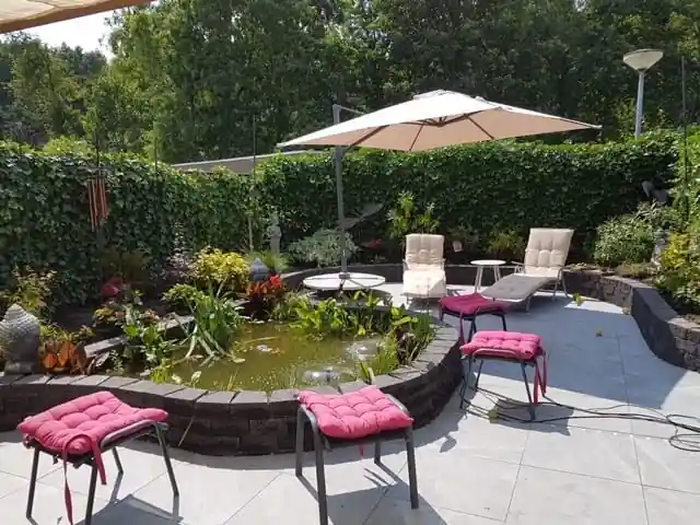 Ronde verhoogde vijver van zwarte stapelblokken met waterplanten en waterlelies, omringd door weelderige borders, een klimopwand en een terras met ligstoelen