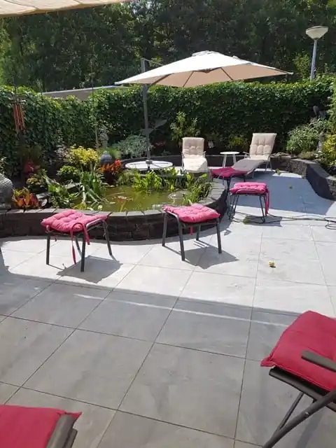 Terras van lichtgrijze tegels met ligstoelen en een zweefparasol naast een verhoogde vijver met waterplanten, omzoomd door een klimopwand en borders