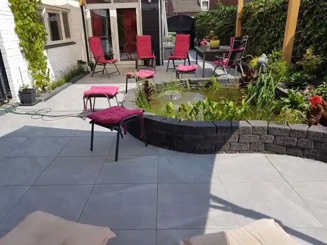 Verhoogde vijver met waterplanten en een fontein, omrand met zwarte stapelblokken, naast een ruim terras van lichtgrijze tegels met tuinstoelen met rode kussens