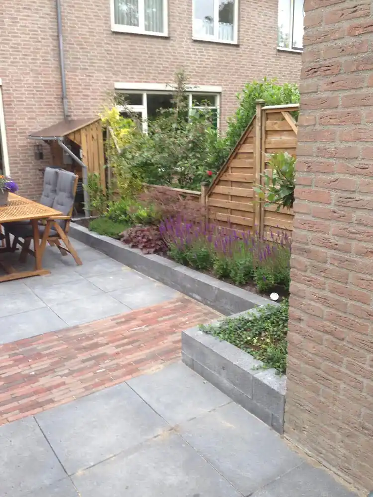 Verhoogde border met paarse salie en heuchera achter een rand van antracieten betonbanden, langs een terras van grijze betontegels met een strook rode klinkers