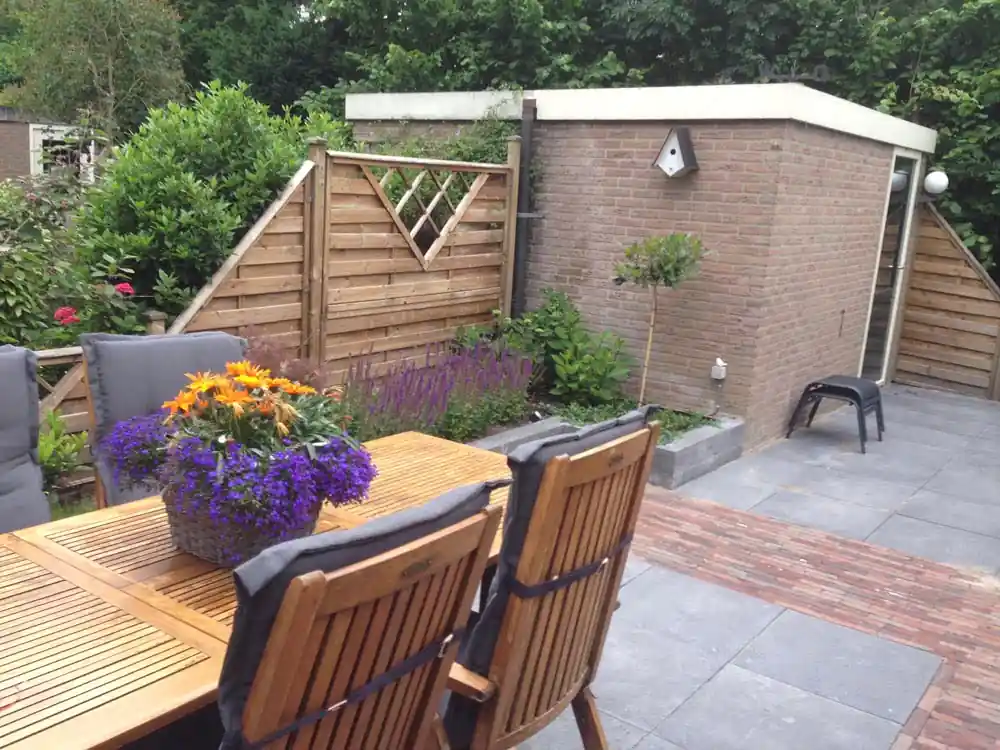Houten eettafel met bloemen op het tegelterras, met zicht op een gemetselde berging, een leiboom op stam en een border met salie langs de schutting