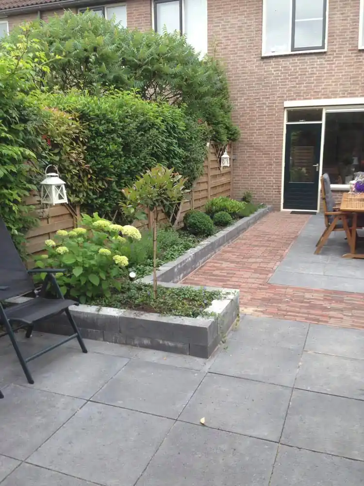 Pad van rode klinkers tussen verhoogde borders met witte hortensia, buxusbollen en een jonge sierboom, leidend naar de achterdeur van de woning