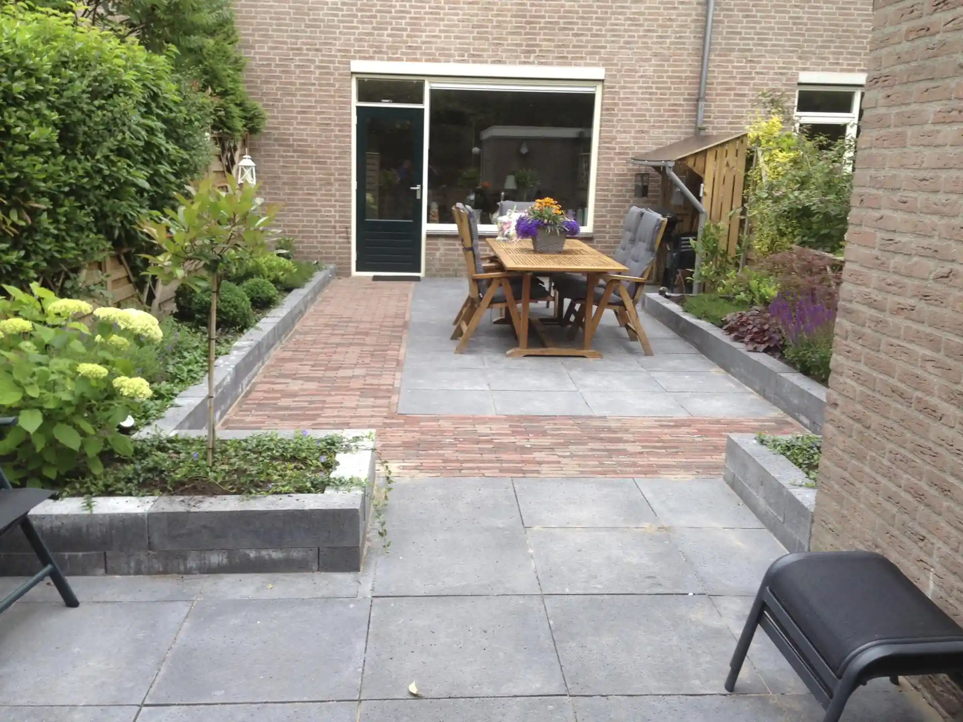 Terras van grote grijze betontegels doorsneden door banen rode klinkers, met een houten eettafel voor de pui en verhoogde borders met hortensia en salie