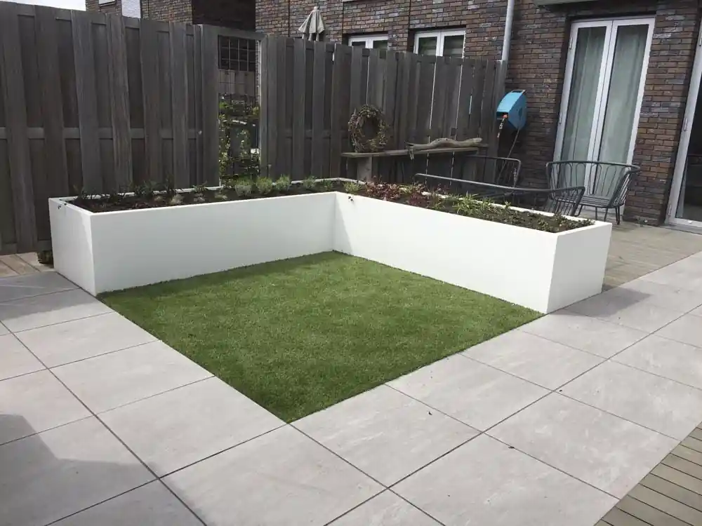 Vierkant veldje kunstgras omsloten door een L-vormige witte plantenbak met verse beplanting, met daaromheen een terras van grote lichtgrijze tegels