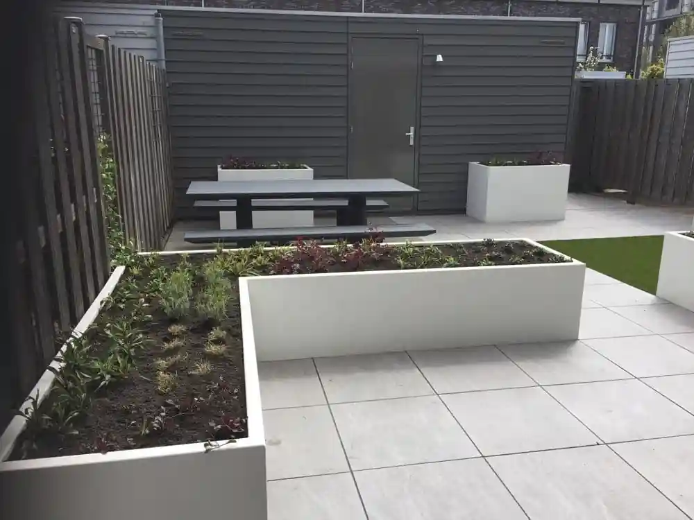 Donkere picknicktafel voor een antracietkleurige berging, omringd door witte plantenbakken met verse beplanting, een tegelterras en een strook kunstgras