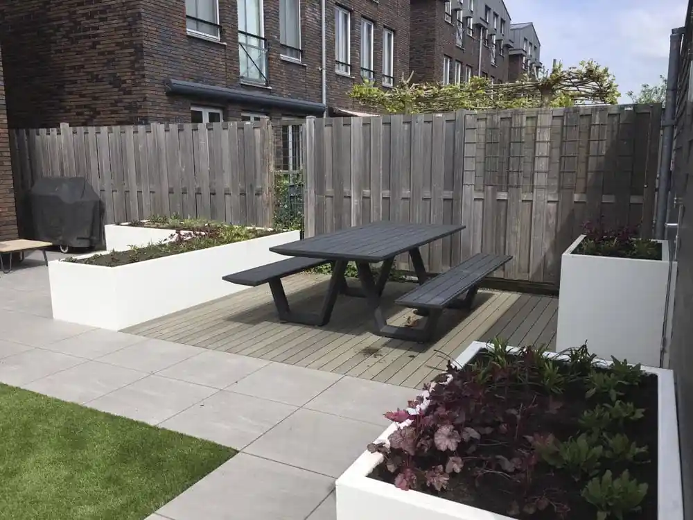 Witte plantenbak met roodbladige heuchera op de voorgrond, met daarachter een picknicktafel op een vlonder en een tegelterras langs de woning