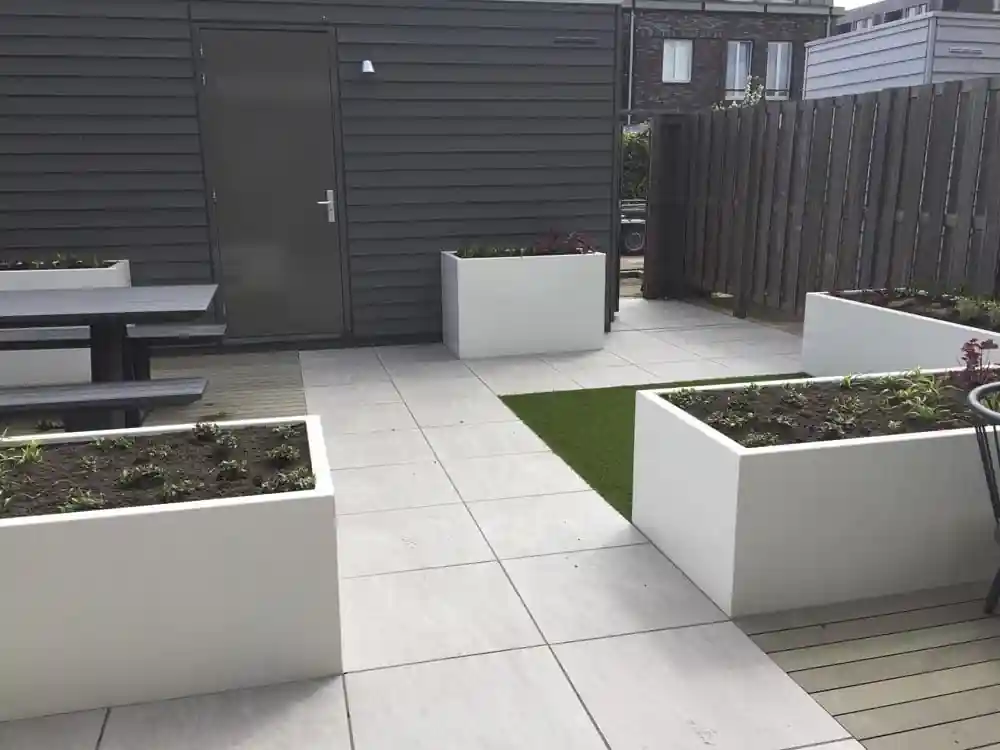 Meerdere witte plantenbakken met verse beplanting op een terras van grote lichtgrijze tegels, met een strook kunstgras en de deur van een donkere berging