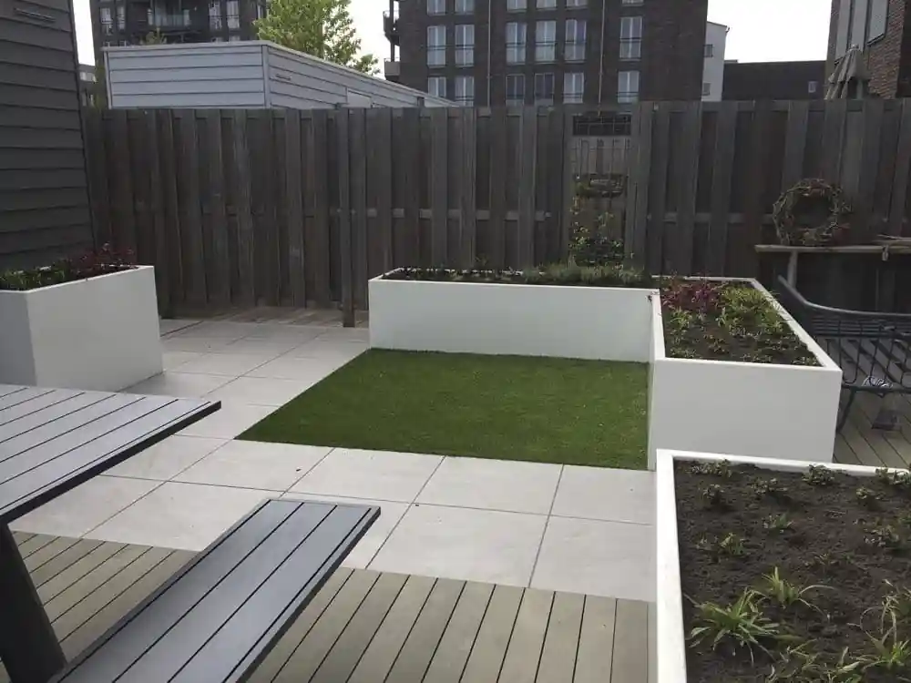Veld kunstgras tussen witte plantenbakken met jonge beplanting, grenzend aan een tegelterras en een vlonder met tuintafel
