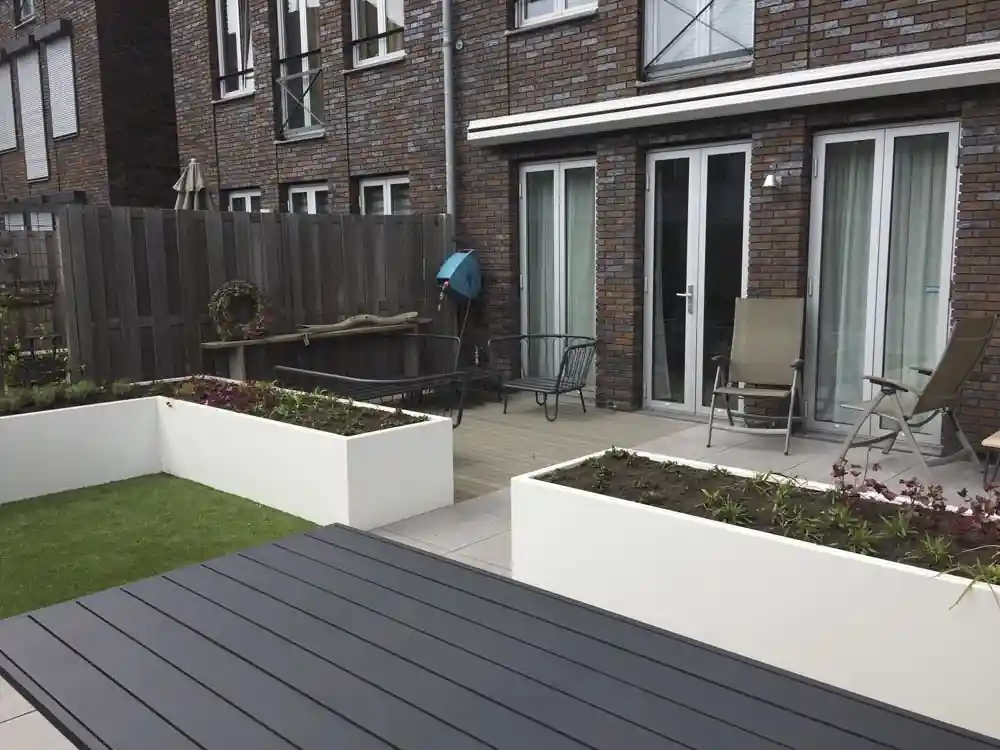 Tegelterras met tuinstoelen voor de openslaande deuren van een bakstenen woning, met witte plantenbakken en een donkere tuintafel op de voorgrond