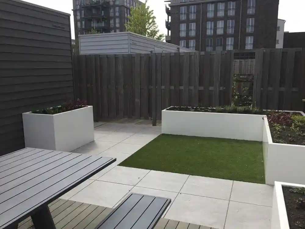 Achtertuin met een tegelterras, een veld kunstgras en meerdere witte plantenbakken, met een houten schutting en appartementengebouwen op de achtergrond