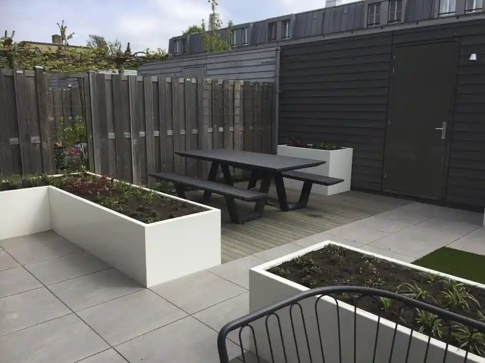 Picknicktafel op een composiet vlonder tussen witte plantenbakken, met rechts een antracietkleurige berging en leibomen boven de schutting