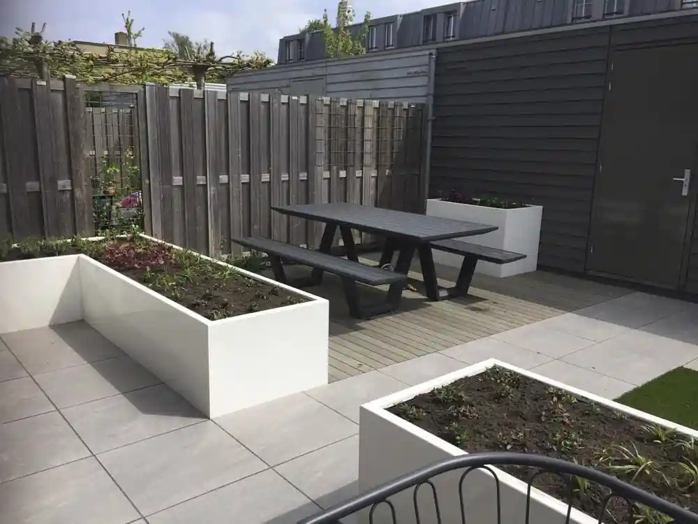 Donkere picknicktafel op een vlonder van composiet planken, met witte plantenbakken eromheen en leibomen boven de houten schutting