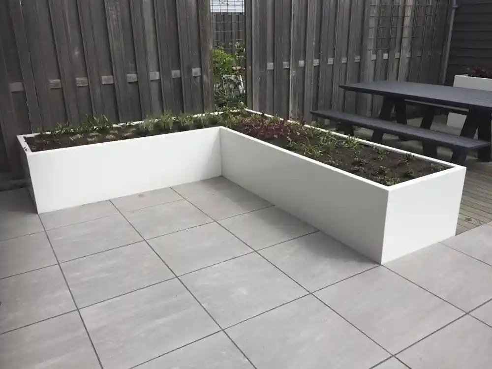 L-vormige witte plantenbak met verse beplanting in de hoek van een terras van grote lichtgrijze tegels, met een donkere picknicktafel en een houten schutting