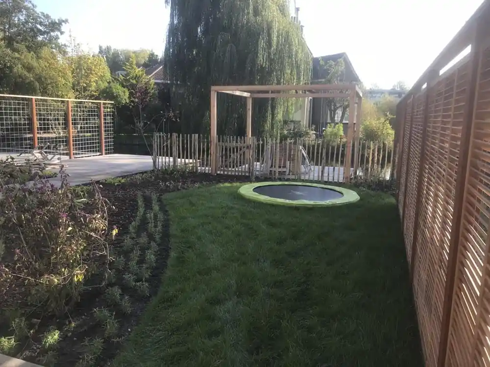 Gazon met een verdiepte trampoline tussen een lattenscherm en een border met vaste planten, met een pergola en de waterkant op de achtergrond
