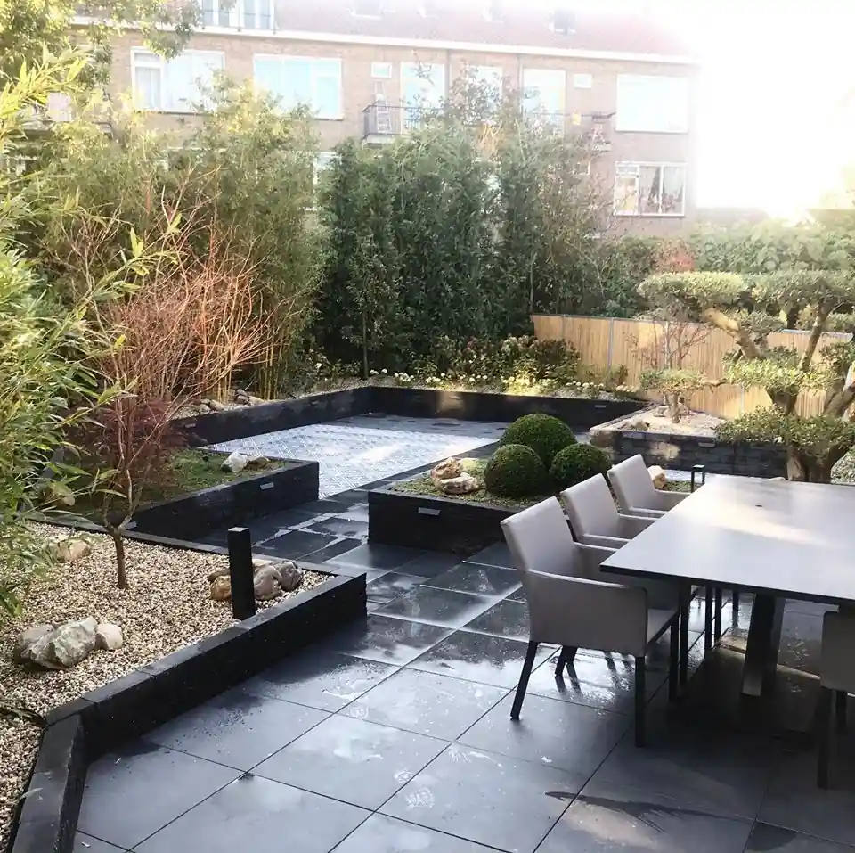 Terras van grote antracieten tegels met een donkere eettafel en lichtgrijze stoelen, met verhoogde borders met buxusbollen, siergrind en zwerfkeien