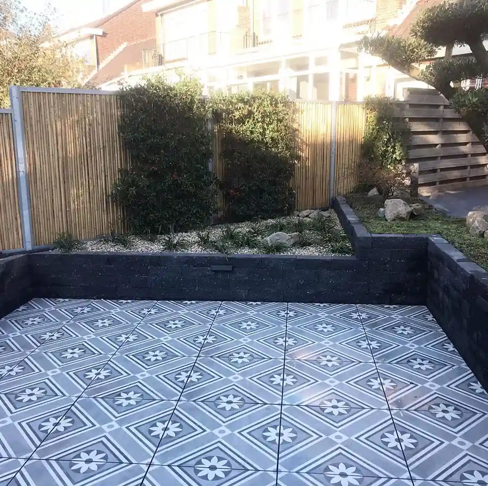 Verdiept terras van witblauwe patroontegels omsloten door keermuren van zwarte stapelblokken, met een bamboeschutting en een border met siergrind en zwerfkeien